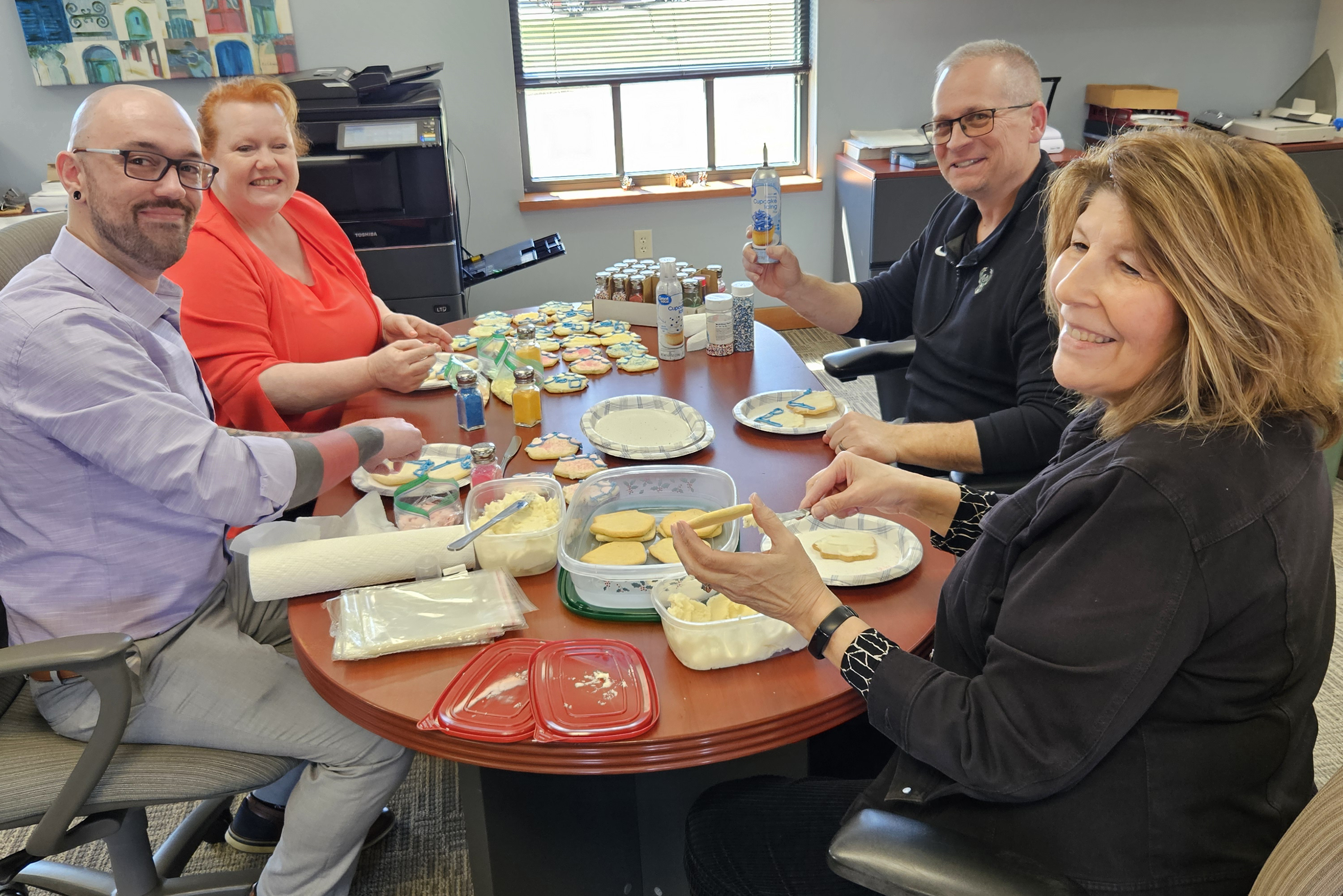 4 people sitting around a table with cookies and cookie decorating supplies covering the table